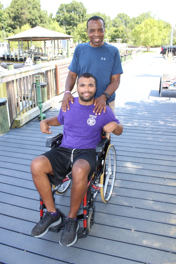 Young adult male guest in wheelchair with male adult caregiver standing behind him. 