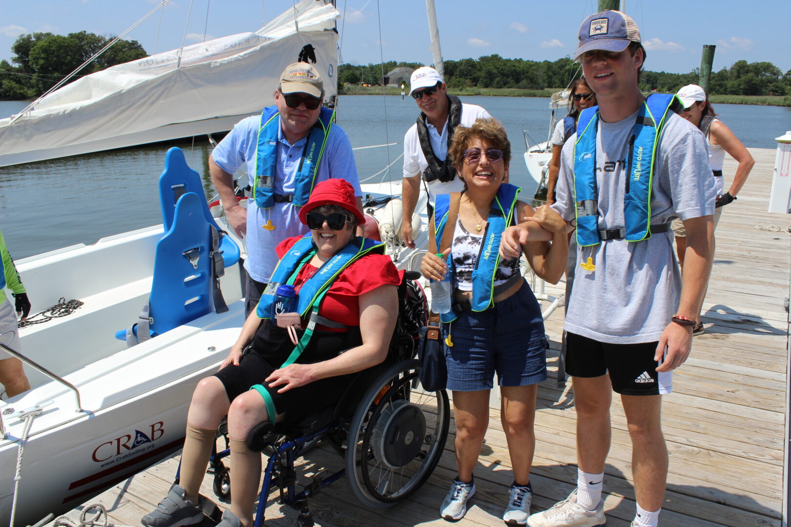 A group photo of 5 individuals, both guests and CRAB volunteers, on the dock next to a CRAB sailboat. 