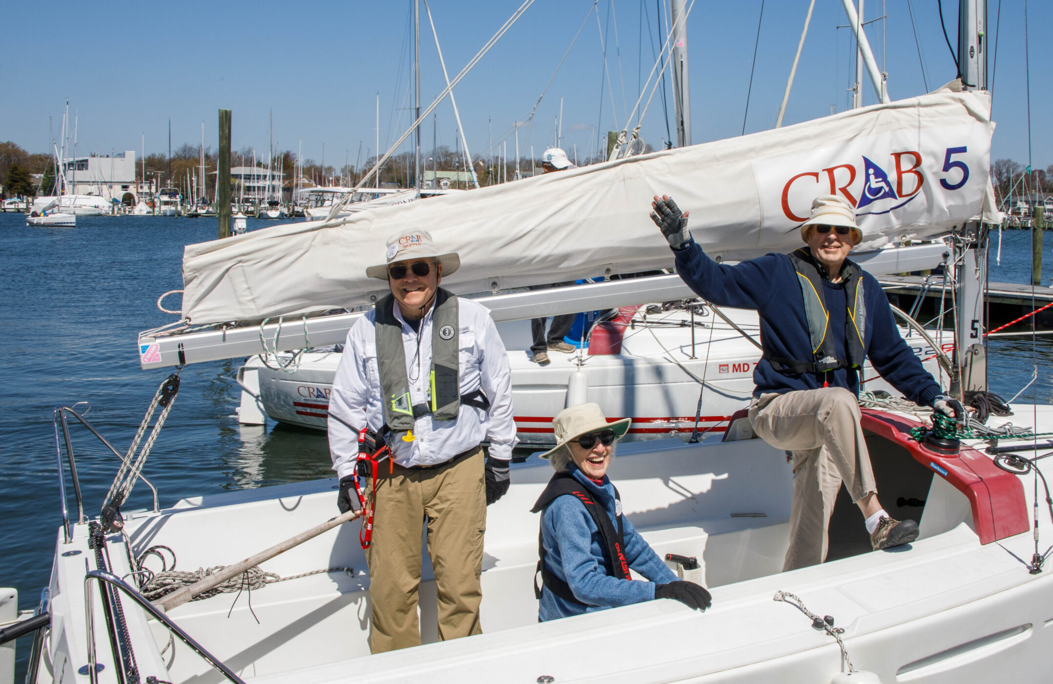 Photo of three CRAB volunteers on a CRAB sailboat, all smiling and one waving to the camera. 