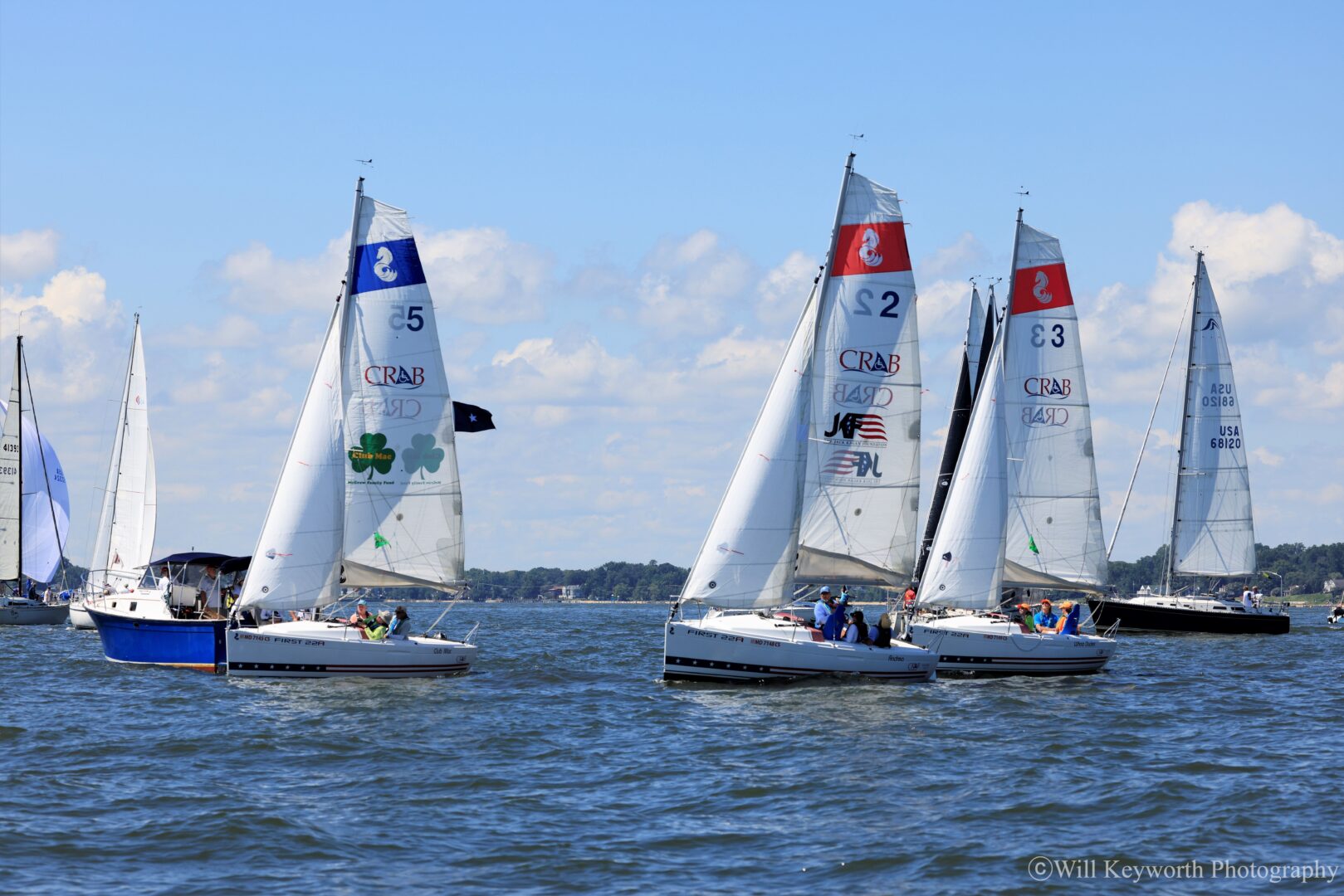 Three CRAB Sailboats under sail in light winds with blue sky. You can see several other boats in the background